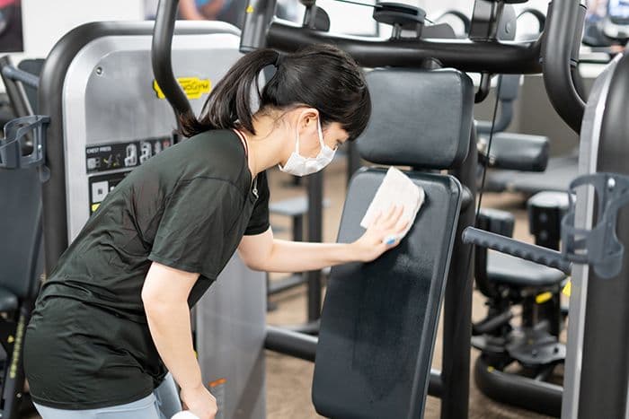 Woman wearing a mask wiping down gym equipment at an apartment fitness center, demonstrating proper fitness etiquette.