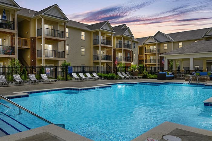 Sparkling outdoor pool with lounge chairs at an Ardent apartment community, featuring multi-story residential buildings at sunset.