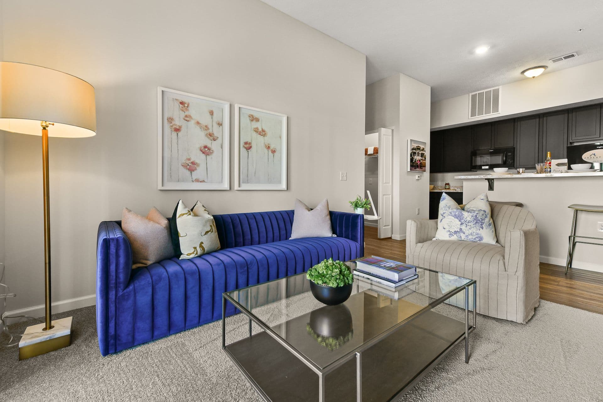 A living room with a blue sofa, gray chair, glass coffee table and kitchen in the background