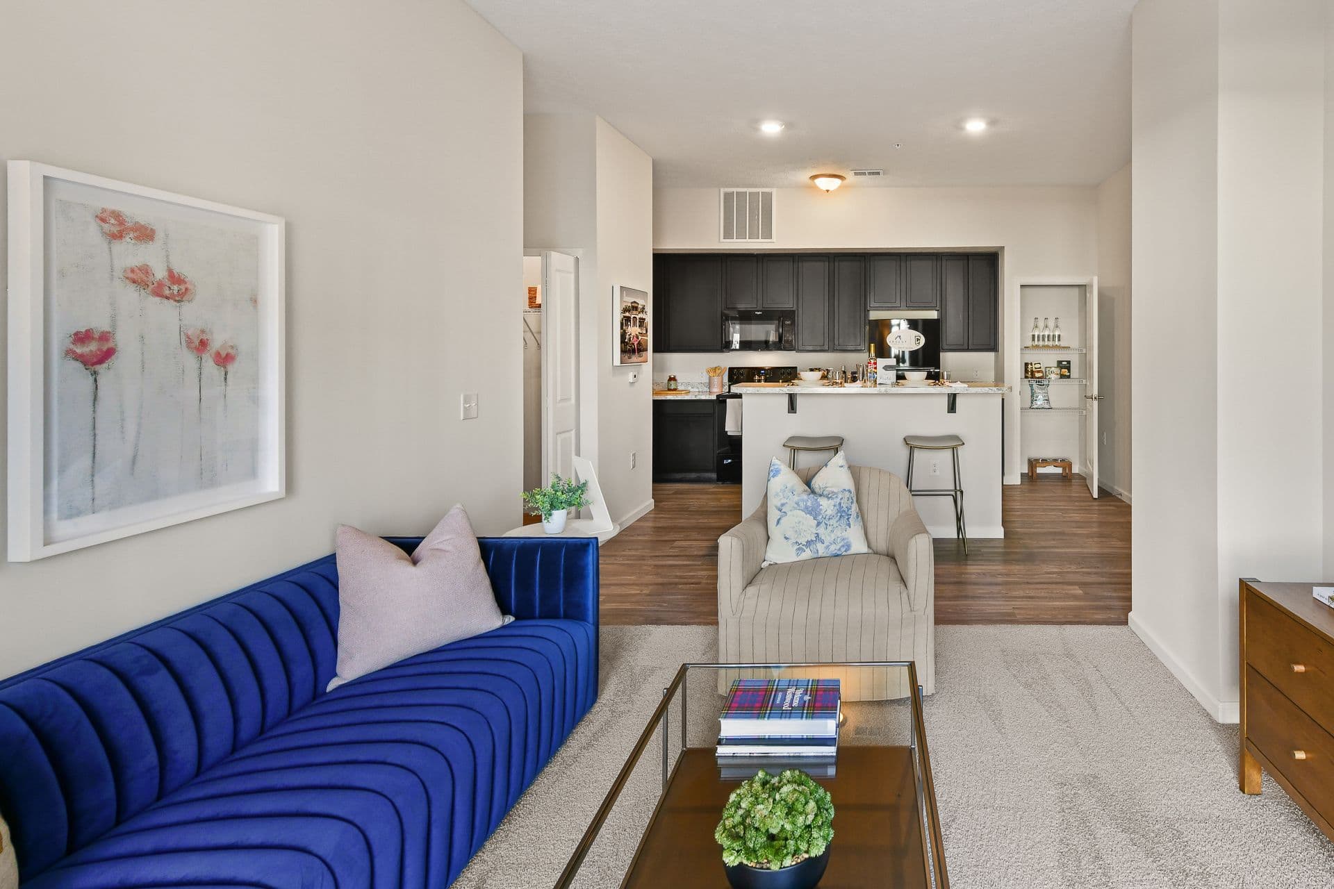 A carpeted living room with a blue sofa, glass table and connecting kitchen