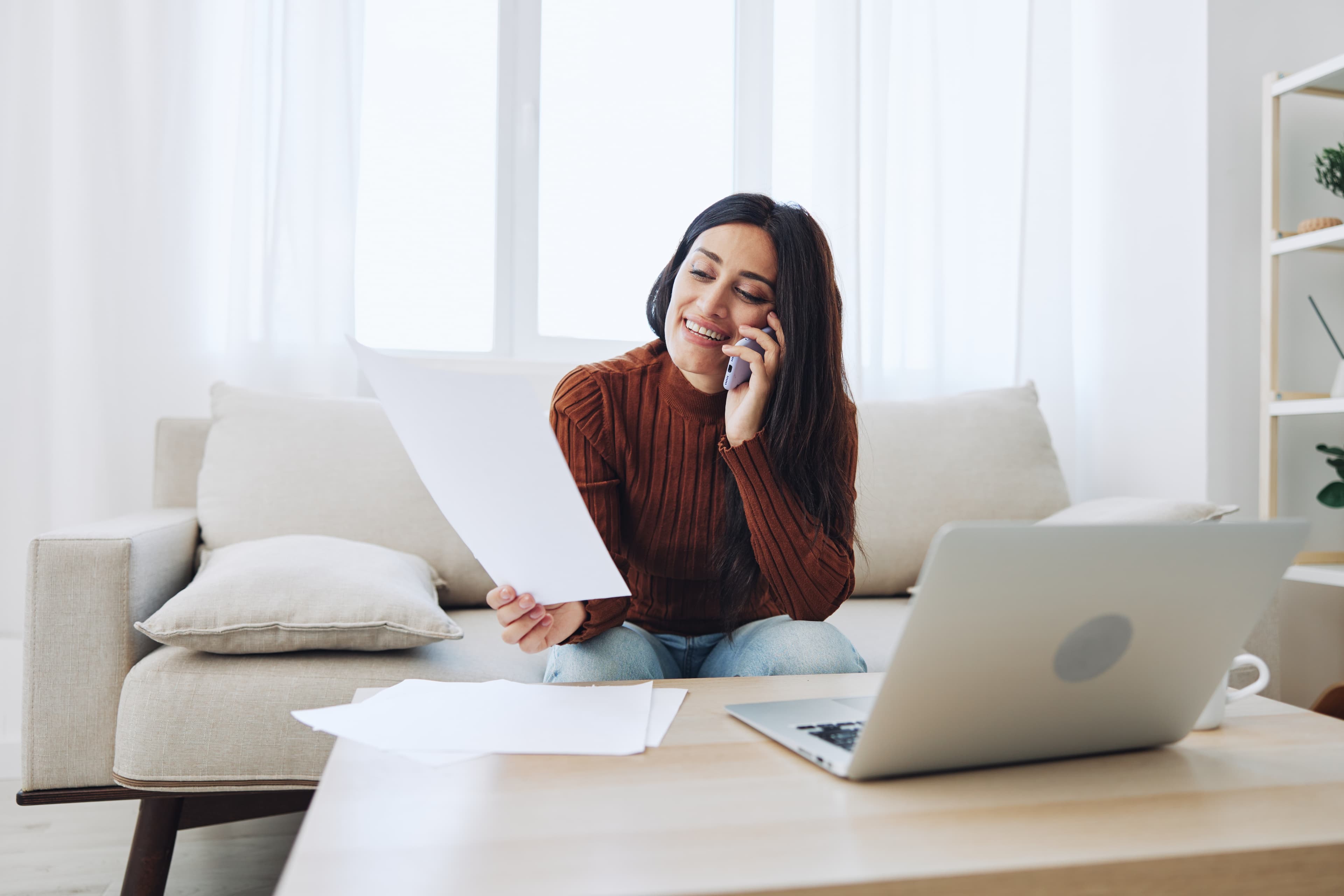 A woman talking on the phone while reviewing paperwork to rent an apartment with a laptop on a table in front of her.